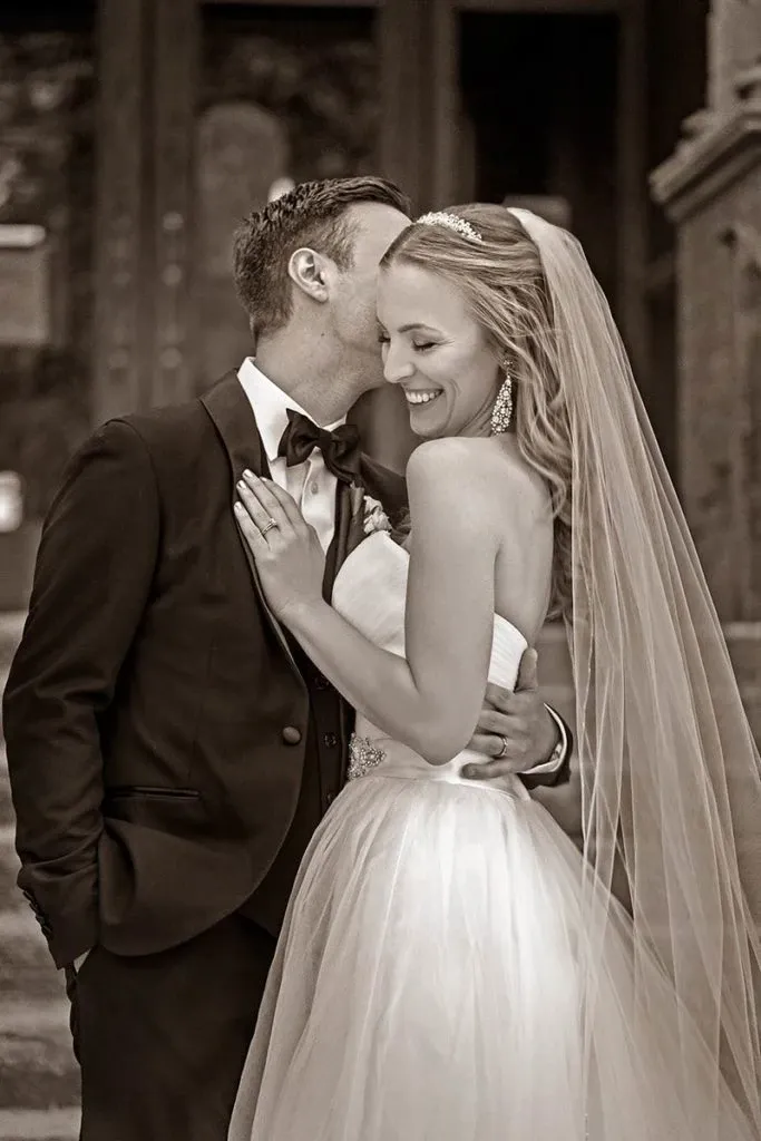 A groom in a classic black tuxedo lovingly kisses his smiling bride on the cheek as she embraces him. She wears a beautiful strapless wedding gown with a delicate veil flowing down her back, complemented by elegant drop earrings and a sparkling headpiece. The background hints at an ornate, historic setting, adding a touch of timeless romance to the scene. Captured by Studio Omega Photo, this image embodies the joy and intimacy of a wedding day, illustrating their expertise in wedding photography and showcased on Mariage Elegant, Canada.