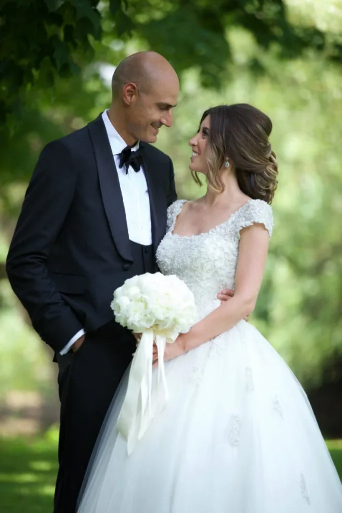 A joyful bride and groom stand closely together in an outdoor setting, surrounded by lush greenery. The groom is wearing a classic black tuxedo with a bow tie, while the bride is dressed in an elegant white gown with intricate lace detailing and short sleeves. She holds a delicate bouquet of white flowers tied with satin ribbons. Their expressions convey happiness and intimacy, capturing a perfect wedding moment. This image, showcasing the work of Studio Omega Photo, is part of the wedding photography category on Mariage Elegant, Canada.