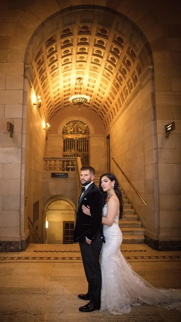 A bride and groom stand elegantly in a grand, historic interior with an ornate ceiling and dramatic lighting, captured by Studio Omega Photo. The bride wears a lace wedding gown and the groom dons a dark suit, both embodying classic wedding style. This image showcases the artistry of Studio Omega Photo in the Wedding Photography category, highlighting a sophisticated and timeless moment in the setting of Mariage Elegant, Canada.