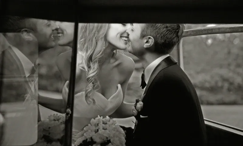 A black and white photograph captured by Studio Omega Photo, specializing in wedding photography, showcases an intimate moment between a newlywed couple. Inside a vintage car, the bride and groom lean in for a tender kiss. The bride is wearing an elegant strapless wedding gown, while the groom is dressed in a classic tuxedo with a bow tie. In the foreground, a bouquet of flowers rests gently, adding a romantic touch to the scene. This heartfelt image beautifully captures the joy and love shared on their special day, exemplifying the artistry of Studio Omega Photo in wedding photography.