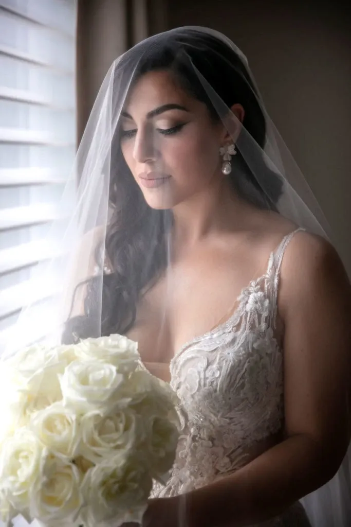 A bride stands gracefully by a softly lit window, holding a bouquet of white roses. She wears a detailed lace wedding gown and a sheer veil that gently frames her face, highlighting her elegant earrings. The reflections in the window cast a warm glow, adding to the romantic ambiance of the moment. This image exemplifies the exceptional work of Studio Omega Photo, a vendor specializing in wedding photography, capturing the beauty and emotion of a bride on her special day for Mariage Elegant in Canada.