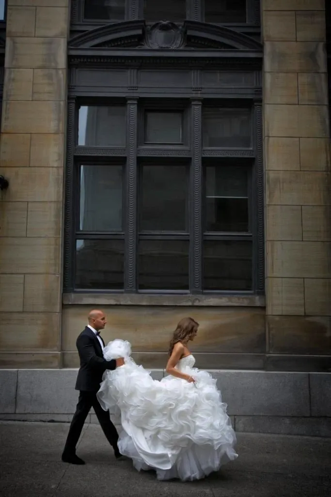A bride and groom walk side by side in front of a grand, historic building with large windows and ornate stone detailing. The groom, dressed in a classic black tuxedo, assists the bride by holding up the train of her voluminous, flowing white wedding gown. The elegant setting and their formal attire highlight the significance of the occasion. This image captures the sophisticated and timeless essence of wedding photography by Studio Omega Photo, featured in the Wedding Photography category on Mariage Elegant, Canada.