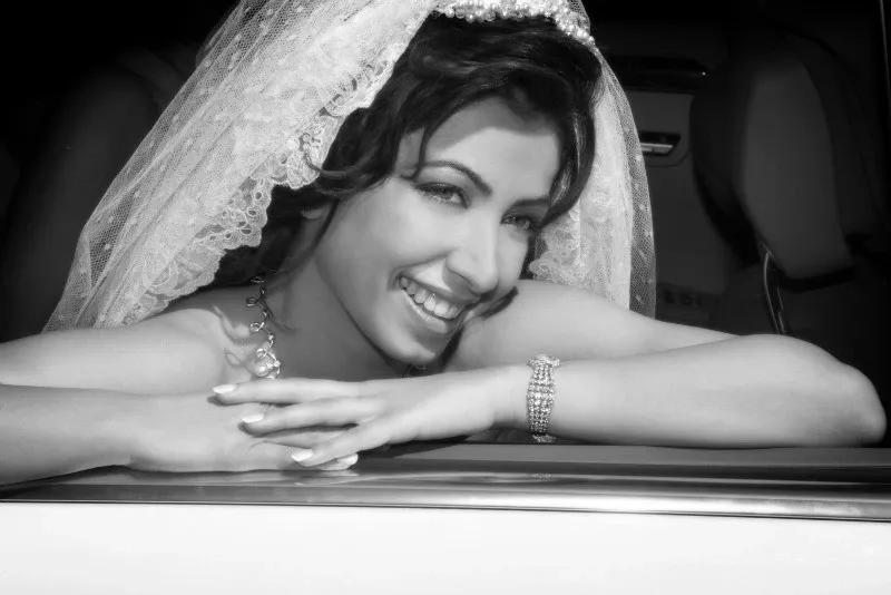 A bride, captured by Studio Omega Photo in the wedding photography category, is leaning out of a car window, exuding happiness. She is smiling brightly, showcasing elegant makeup and intricate jewelry, including a delicate necklace and bracelet. Her veil is intricately designed, adding a classic touch to her bridal attire. This image beautifully represents the joy and elegance of a wedding day, highlighting the exceptional work of a skilled wedding photographer.