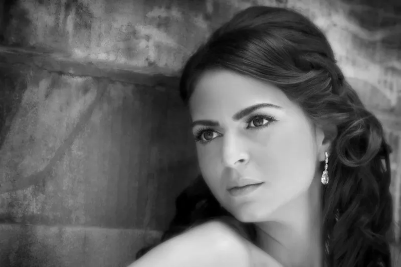 This black and white image captures an elegant bridal portrait by Studio Omega Photo, renowned in the wedding photography category. The bride is depicted leaning against a textured stone wall, her expression contemplative. Her hair is styled in soft, cascading waves, adorned with delicate earrings that add subtle sophistication. The photograph highlights the graceful details of her wedding look, showcasing Studio Omega Photo's expertise in capturing timeless wedding moments for Mariage Elegant in Canada.