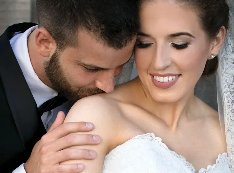 A bride and groom share an intimate moment, captured by Studio Omega Photo, featured in the Wedding Photography category on Mariage Elegant, Canada. The groom, dressed in a classic black suit, lovingly holds the bride's shoulder while gently leaning in. The bride, wearing a beautiful lace wedding dress and delicate veil, smiles softly, exemplifying joy and elegance on their wedding day.