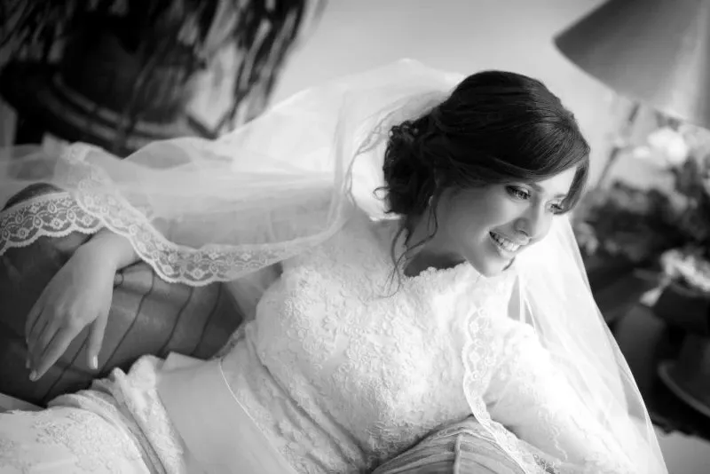 A bride in a lace wedding gown and veil smiles while gracefully leaning on a sofa, captured in black and white by Studio Omega Photo. Her elegant attire is complemented by intricate lace details on both her dress and veil, emphasizing the artistry of wedding photography. The soft lighting creates a romantic and timeless ambiance, perfect for a wedding day moment. This image highlights the expertise of Studio Omega Photo in the wedding photography category, showcasing their ability to capture intimate and beautiful wedding moments.
