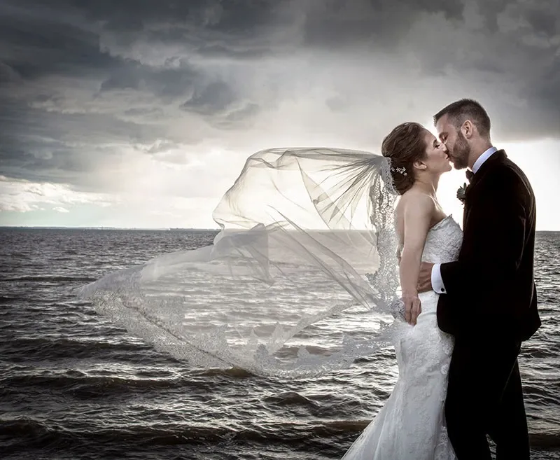 A newlywed couple shares a romantic kiss by the lakeside, captured by Studio Omega Photo under dramatic, cloud-filled skies. The bride's elegant veil flows gracefully in the wind, embodying the essence of wedding photography. The groom, in a classic tuxedo, embraces the bride, who is stunning in a strapless lace gown. This image perfectly showcases Studio Omega Photo's ability to blend natural beauty with intimate moments, ideal for Mariage Elegant's wedding photography category in Canada.