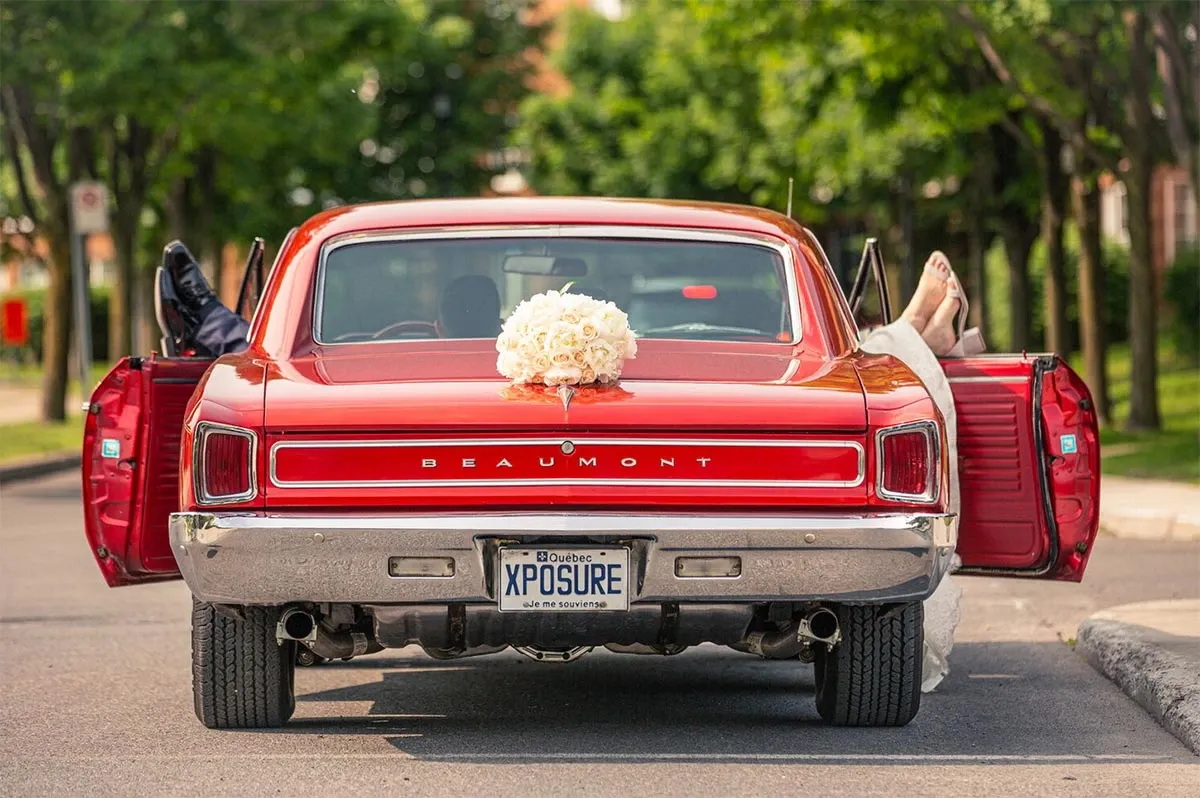 This image by Xposure Photographie, a featured vendor in the Wedding Photography category at Mariage Elegant, Canada, captures a creative and playful scene from a wedding. It shows a classic red Beaumont car with its doors open, parked on a tree-lined street. At the back of the car, a bridal bouquet is elegantly placed on the trunk lid. Two people, likely the newlyweds, are playfully lounging with their feet sticking out of each open car door, one in a black suit and the other in a white wedding dress. The license plate humorously reads 'XPOSURE,' adding a personalized touch to this memorable wedding moment.