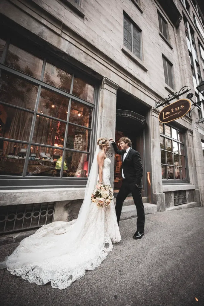 The image features a newlywed couple posing elegantly in front of a vintage building with large windows, captured by Xposure Photographie, a vendor in the Wedding Photography category. The bride is wearing a long, flowing lace dress with a train, and holding a bouquet of flowers, while the groom is dressed in a classic black suit. The setting appears to be urban, adding a sophisticated backdrop to this romantic scene. This photograph is part of the wedding services offered by Mariage Elegant in Canada.
