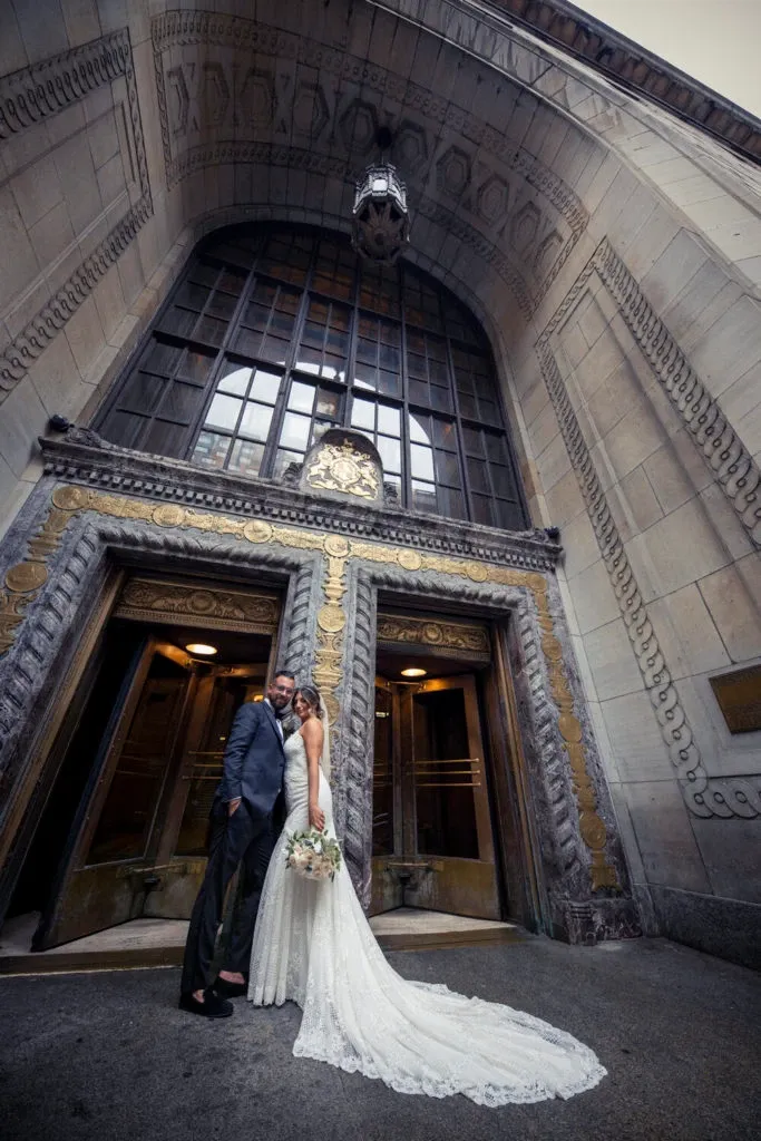 A newlywed couple stands lovingly in front of an ornate, historical building entrance with tall, decorative wooden doors and intricate stone carvings. The bride, wearing an elegant, lace wedding gown and holding a bouquet of pastel flowers, gazes into the eyes of her groom, who is dressed in a tailored suit. This romantic moment is captured by Xposure Photographie, a wedding photography vendor featured on Mariage Elegant, Canada. The setting's grand architectural details provide a classic backdrop, enhancing the timeless feel of this wedding portrait.