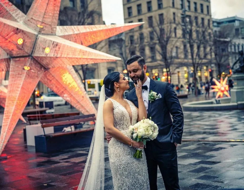This image showcases a loving couple on their wedding day, captured by Xposure Photographie, a vendor in the Wedding Photography category. The bride, wearing an elegant, sparkling gown and a long veil, gently holds a bouquet of white flowers, while the groom, dressed in a formal dark suit with a white boutonniere, lovingly touches her chin. They are standing in an urban setting with large illuminated star decorations, suggesting a festive or winter theme. The scene reflects a moment of intimacy and joy amidst the backdrop of a beautifully decorated cityscape.