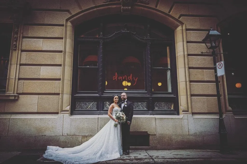 A bride and groom stand elegantly in front of a historic stone building in an urban setting. The bride is wearing a flowing white lace wedding gown with a long train, holding a lush bouquet of flowers. The groom is dressed in a classic black tuxedo with a bow tie. Above them, a large window with dark trim is visible, featuring the word 'dandy' in red letters. The scene is slightly overcast, adding a romantic ambiance to the moment. This image is a part of the wedding photography portfolio from Xposure Photographie, showcased on the Mariage Elegant, Canada website under the Wedding Photography category.