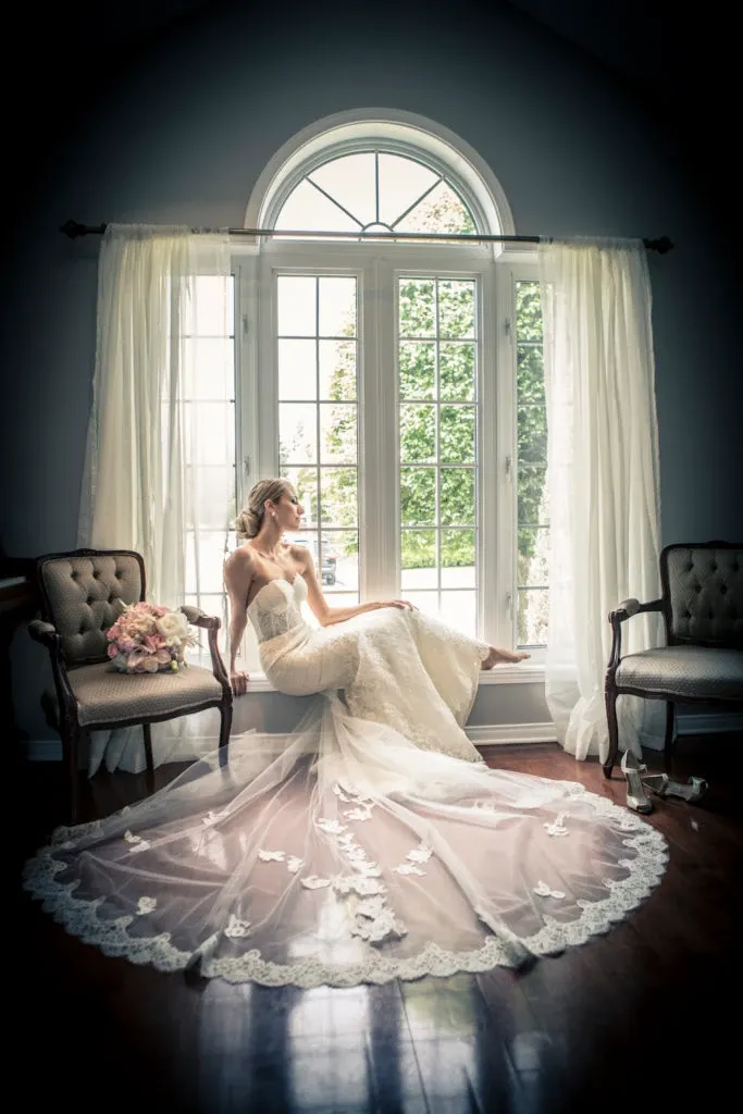 A bride in a delicate, lace wedding gown sits gracefully on the windowsill of a large, arched window, surrounded by flowing white curtains. Her long veil, adorned with intricate floral details, cascades onto the polished wooden floor. Two elegant chairs frame the scene on either side. A bouquet of soft pink and white flowers rests on one chair, adding a touch of color. This image, captured by Xposure Photographie, perfectly encapsulates the romantic essence of wedding photography, showcasing their expertise in creating timeless, elegant bridal portraits for Mariage Elegant, Canada.