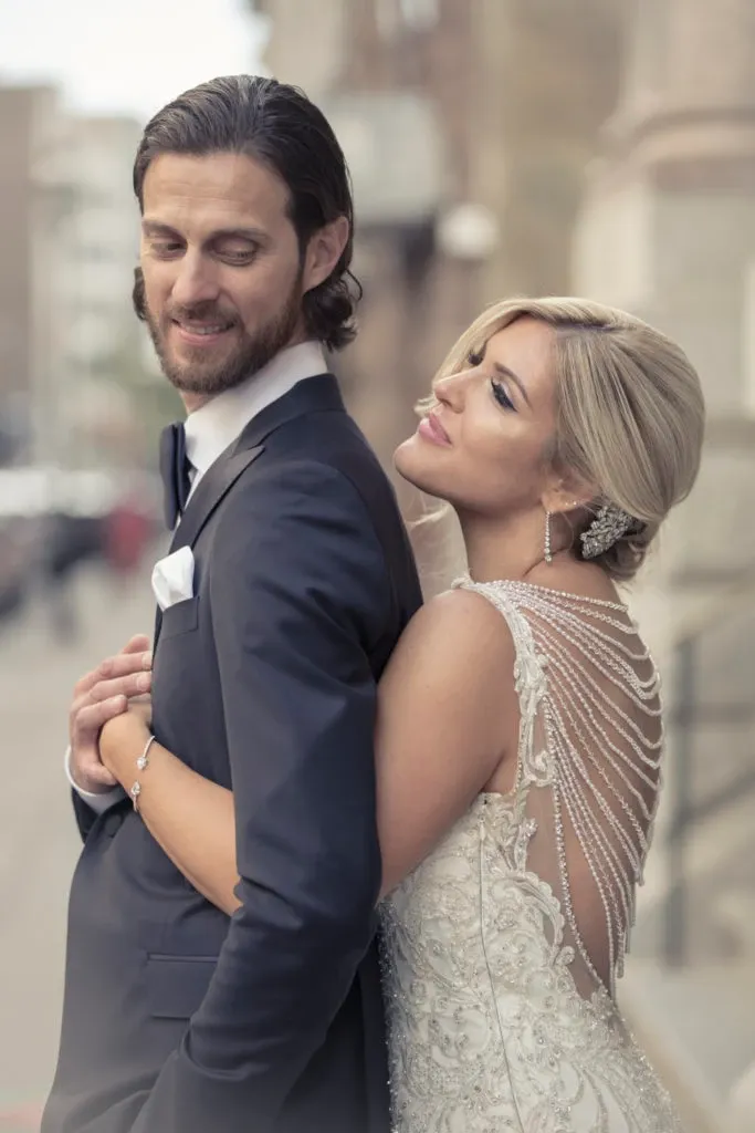A bride and groom are captured in a tender moment on their wedding day by Xposure Photographie, a vendor specializing in wedding photography in Canada for the Mariage Elegant platform. The bride, wearing an intricately designed gown with beaded details and an elegant hairpiece, lovingly embraces the groom from behind as he stands in a dark suit. They are posed outdoors against a blurred urban backdrop, adding a romantic and timeless feel to the image.