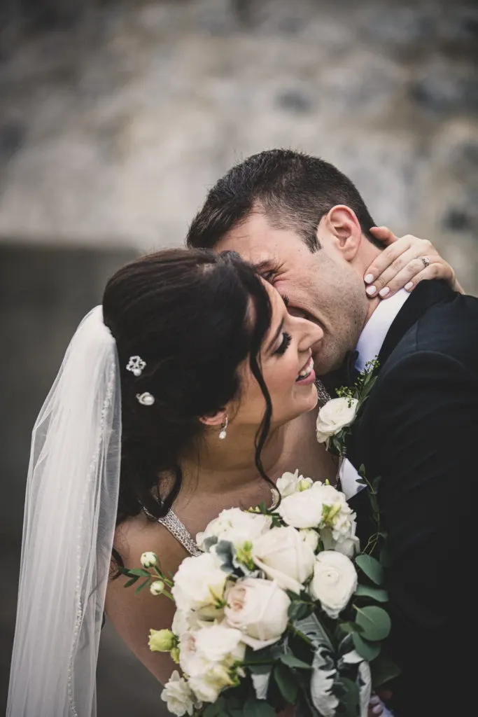 A joyful bride and groom share a tender embrace, captured by Xposure Photographie, a vendor specializing in wedding photography. The bride, adorned with a delicate white veil and elegant hair accessories, holds a beautiful bouquet of white roses and greenery. The groom, wearing a classic black suit, leans into her with a warm smile, epitomizing the happiness of a wedding day. This intimate moment showcases the couple's love and is a great example of the artistry offered by Xposure Photographie in capturing timeless wedding memories.