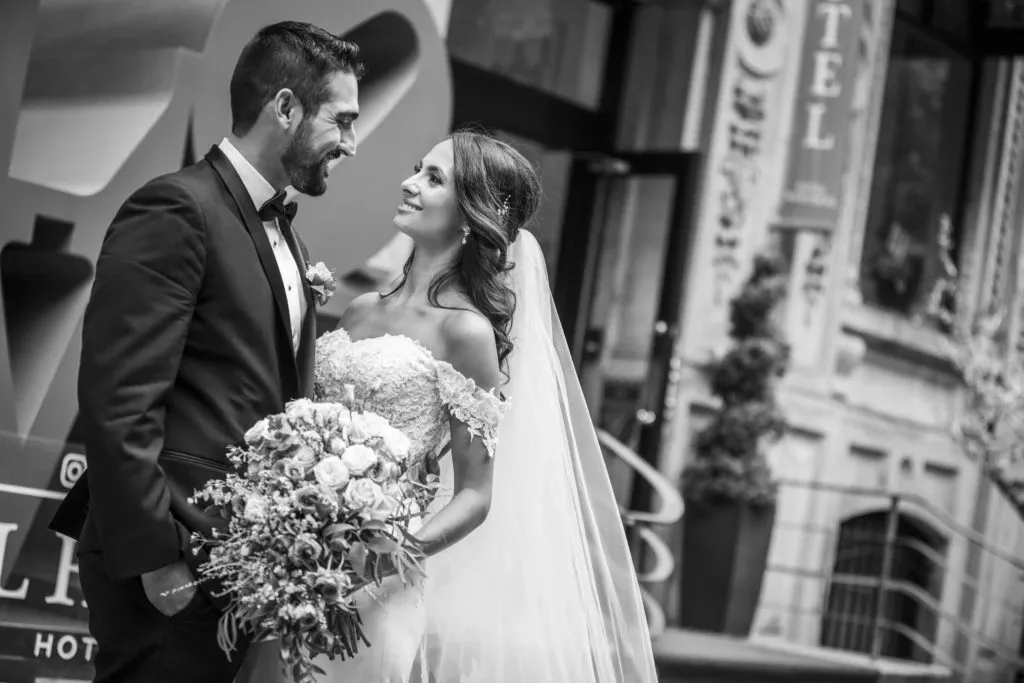 In this elegant black and white wedding photograph by Xposure Photographie, a joyful bride and groom share a loving gaze outside a stylish venue. The bride is wearing an off-shoulder lace wedding gown and a flowing veil while holding a lush bouquet of flowers. The groom is dressed in a sharp tuxedo. Their surroundings hint at a sophisticated city location, with a backdrop of chic architectural details and hotel signage. This image beautifully captures an intimate moment in the context of wedding photography, showcasing Xposure Photographie's skill in creating timeless wedding memories.