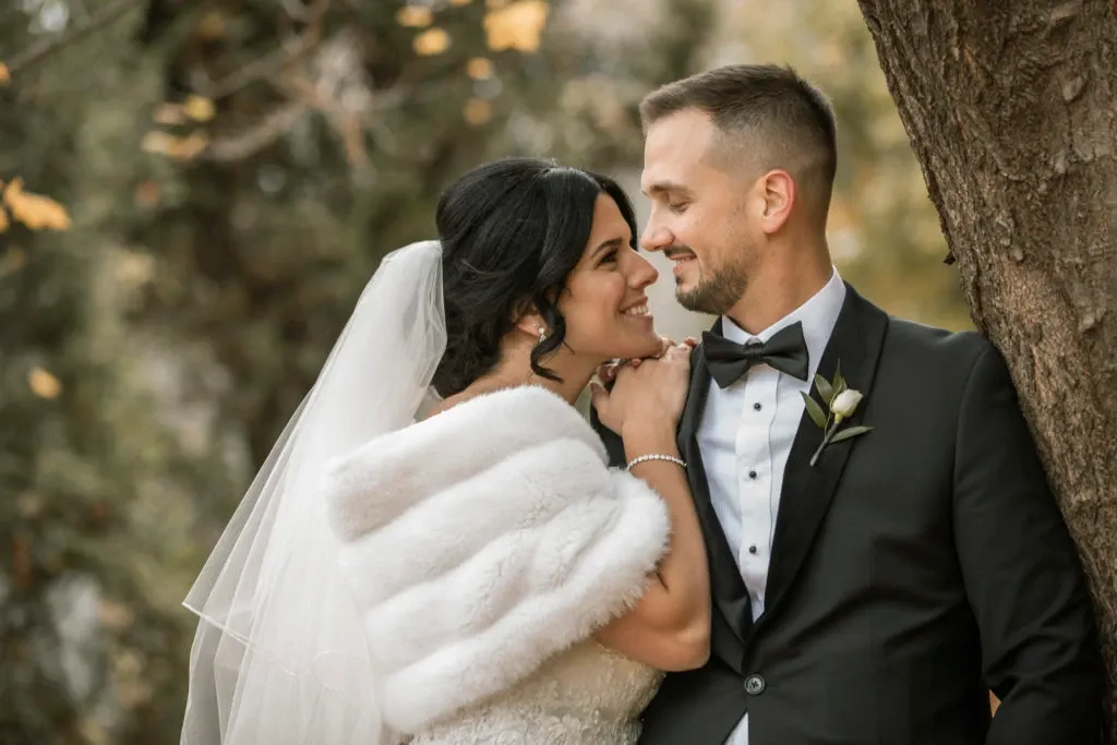 This image showcases a joyful couple on their wedding day, captured beautifully by Xposure Photographie, part of the Wedding Photography category on Mariage Elegant, Canada. The bride is dressed in an elegant white gown with a fur wrap and a veil, gently leaning against a tree as she smiles warmly at the groom. The groom is wearing a classic black tuxedo with a white shirt and black bow tie, looking affectionately at the bride. The background features soft, blurred greenery, adding a natural charm to this romantic outdoor moment.