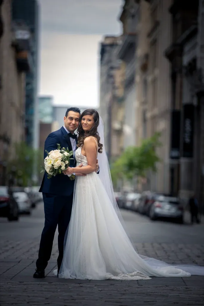 A bride and groom pose together in an urban setting for their wedding photoshoot, captured by Xposure Photographie, under the wedding photography category at Mariage Elegant, Canada. The bride is wearing a flowing white gown with intricate lace details and a long veil, while the groom is dressed in a classic dark suit. The bride holds a bouquet of white and blush roses. They are standing on a cobblestone street, with historic buildings and blurred city architecture in the background, creating a romantic and timeless atmosphere.