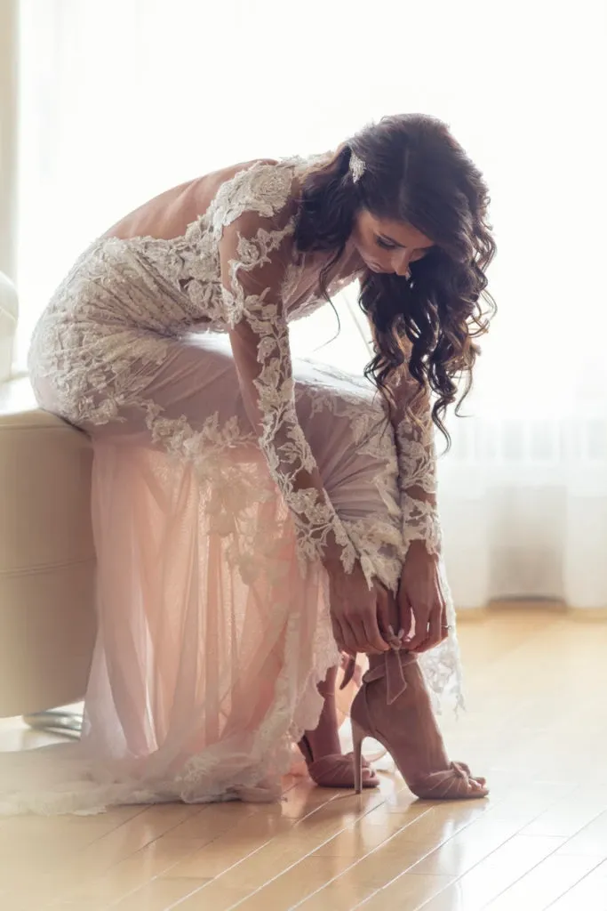 In this image by Xposure Photographie, a bride is captured in a serene moment as she delicately secures her high heel shoe. Dressed in an elegant lace wedding gown, she is seated on a soft, upholstered bench with a glow from natural light streaming through a nearby window. The intricate lace detailing of her dress complements the soft blush tones, embodying a sense of romance and sophistication, characteristic of wedding photography. This image is a beautiful representation of the wedding preparation process, showcasing the bride's elegance and attention to detail.