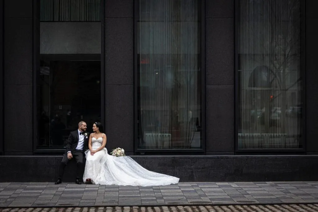 A newlywed couple is sitting closely together outside a modern building, with large windows serving as a backdrop. The bride is dressed in an elegant white wedding gown with a long train, and she holds a bouquet of flowers. The groom wears a classic black tuxedo. They both appear relaxed and content, gazing lovingly at each other. This moment captures the essence of a sophisticated urban wedding. The photograph, taken by Xposure Photographie, highlights their expertise in wedding photography, showcasing a blend of romance and contemporary style in the heart of a cityscape.