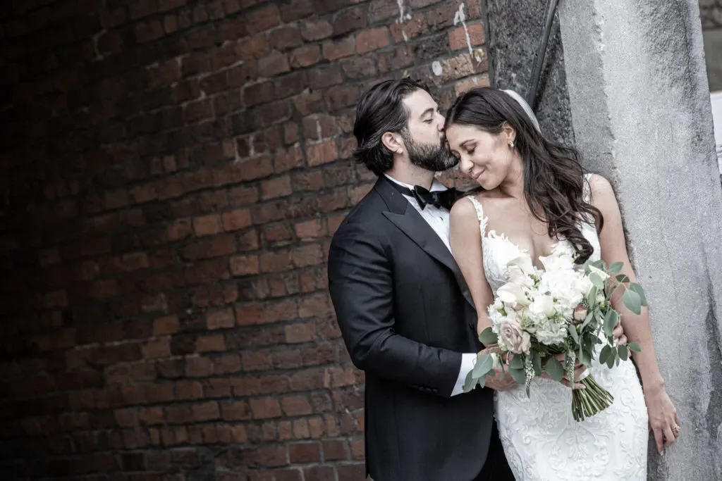A bride and groom are captured in an intimate moment by Xposure Photographie, under the wedding photography category. The groom, dressed in a classic black tuxedo with a bow tie, gently kisses the bride's forehead as she smiles softly, holding a large bouquet of white and blush flowers with green leaves. The bride is wearing an elegant, lace wedding gown, and her long, dark hair cascades down her shoulders, accentuated by a delicate headpiece. The couple stands against a rustic brick wall and a concrete pillar, creating a romantic and timeless wedding scene.