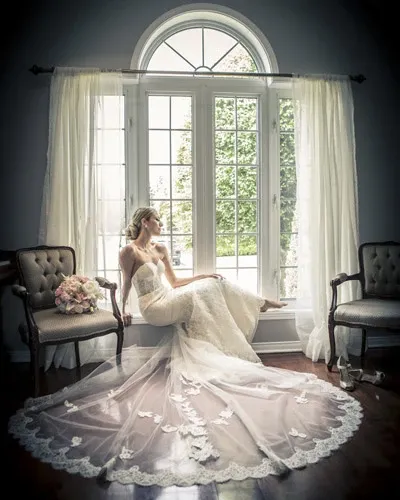 A bride in an elegant lace wedding gown sits gracefully on a window ledge, with the train of her dress cascading onto the wooden floor, adorned with intricate floral patterns. Soft natural light filters through the large, arched window behind her, creating a serene and romantic atmosphere. Two vintage chairs flank the window, one displaying a delicate bouquet of pale pink roses. This image is a stunning example of Xposure Photographie's work in the wedding photography category, capturing the beauty and elegance of a bridal portrait for Mariage Elegant in Canada.