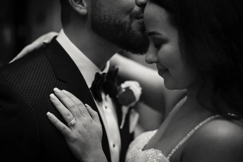 A black and white image showcasing a tender moment between a bride and groom. The bride gently rests her hand on the groom's chest, highlighting her elegant engagement ring. She smiles softly with her eyes closed as the groom tenderly kisses her forehead. The groom is wearing a textured tuxedo jacket and bow tie, with a boutonniere pinned to his lapel. This intimate scene is captured by Xposure Photographie, a vendor specializing in wedding photography for Mariage Elegant in Canada.