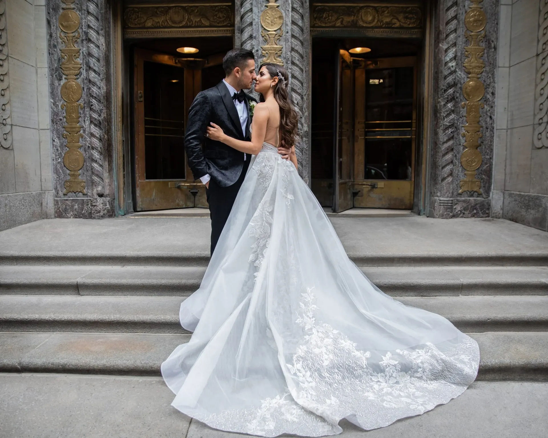 In this image from the wedding photography category by Foto Studio Franca Sorrenti, a bride and groom share an intimate moment on the steps of an elegantly designed stone building. The bride is wearing a stunning white gown with intricate lace detailing and a long, flowing train, while the groom is dressed in a classic black tuxedo. The backdrop features ornate architectural elements that add to the grandeur of the scene, capturing the essence of a sophisticated wedding venue in Canada. This photograph exemplifies the artistic quality and romantic ambiance offered by Foto Studio Franca Sorrenti.