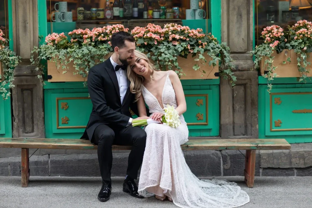 In this charming image captured by Foto Studio Franca Sorrenti, a couple is pictured sitting on a wooden bench in front of a vibrant storefront adorned with potted flowers, highlighting their role in the wedding photography category. The groom, dressed in a classic black tuxedo, lovingly embraces the bride, who is wearing an elegant white lace wedding gown. The bride holds a delicate bouquet of white flowers, and both appear blissfully content, capturing a romantic and intimate moment. This photograph is an exemplary showcase of wedding photography craftsmanship, perfect for a wedding planning setting on Mariage Elegant, Canada.