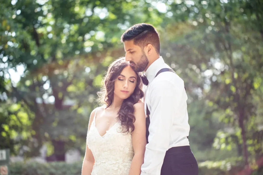 A newlywed couple stands closely together in a serene outdoor setting, captured by Foto Studio Franca Sorrenti in the Wedding Photography category for Mariage Elegant, Canada. The bride, wearing a lace wedding dress with delicate straps, gently rests her head on the groom's shoulder. The groom, dressed in a crisp white shirt and black suspenders, tenderly embraces her. Sunlight filters through the lush green trees in the background, creating a soft and romantic atmosphere.
