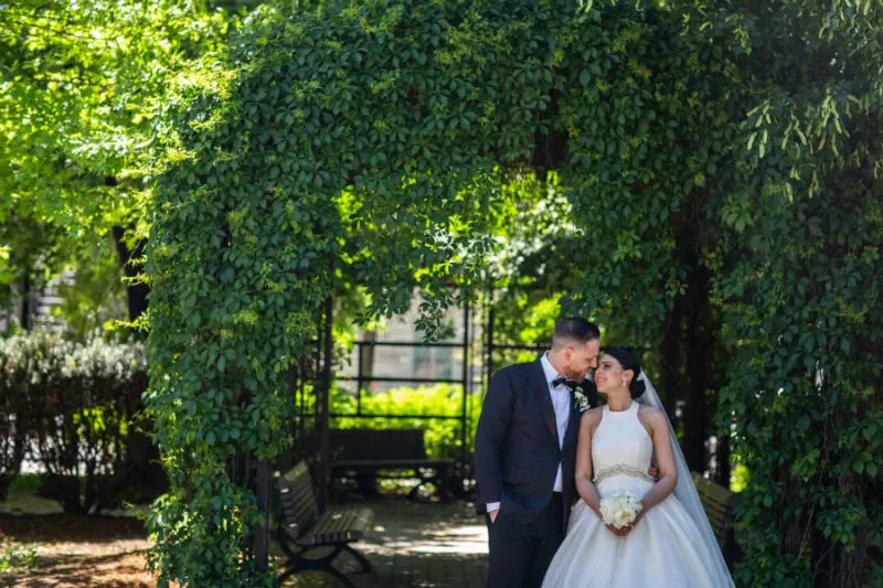 A bride and groom share a tender moment under a lush, green archway in a serene garden setting. The bride is elegantly dressed in a flowing white gown and holds a bouquet of flowers, while the groom looks sharp in a classic dark suit. Sunlight filters through the leaves, creating a romantic atmosphere. This image captures the expertise of Foto Studio Franca Sorrenti in the Wedding Photography category, highlighting a perfect moment of joy and love suitable for any wedding celebration.