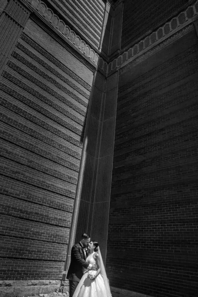 A newlywed couple stands closely together at the base of an imposing brick building, with architectural elements casting dramatic shadows around them. The bride, wearing an elegant white gown with lace detailing, holds a bouquet while gazing lovingly at the groom, who is dressed in a formal dark suit. The towering brick walls provide a striking urban backdrop, adding contrast to the couple's romantic pose. This captivating moment is captured by Foto Studio Franca Sorrenti, a vendor specializing in wedding photography for the Mariage Elegant platform in Canada.