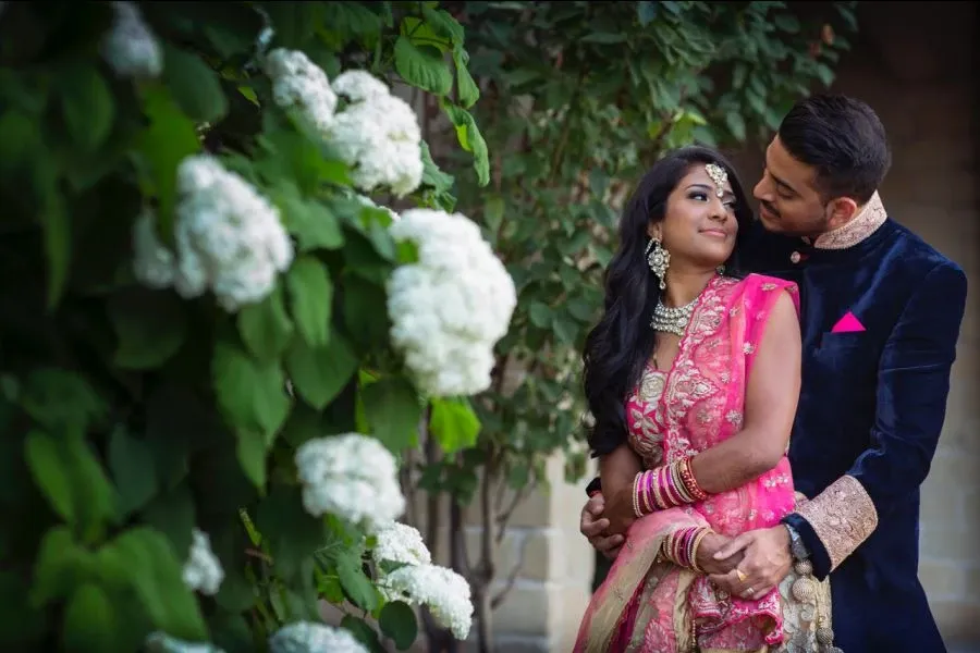 A bride and groom are joyfully embracing in a lush garden, surrounded by blooming white flowers. The bride, adorned in a vibrant pink and gold traditional wedding saree and elegant jewelry, looks lovingly at her groom. The groom is dressed in a classic navy blue sherwani with a pink pocket square, complementing the bride's attire. The serene backdrop and the couple's joyful expressions epitomize the elegance of a wedding celebration. This image is captured by Foto Studio Franca Sorrenti, showcasing their expertise in wedding photography.