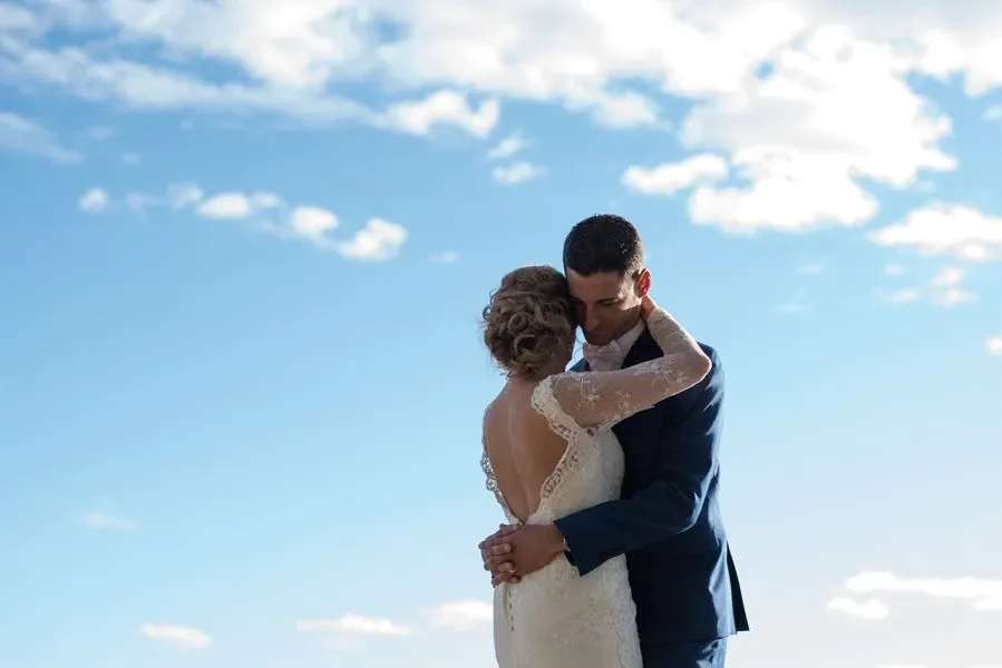 A newlywed couple embraces under a bright blue sky, with soft, scattered clouds providing a serene backdrop. The bride, wearing an elegant lace dress with a low back, has her hair styled in an intricate updo. The groom, in a tailored navy suit, holds her gently. This romantic moment is expertly captured by Foto Studio Franca Sorrenti, a specialist in wedding photography. The image conveys a sense of intimacy and joy, highlighting the vendor's skill in capturing precious wedding day memories.