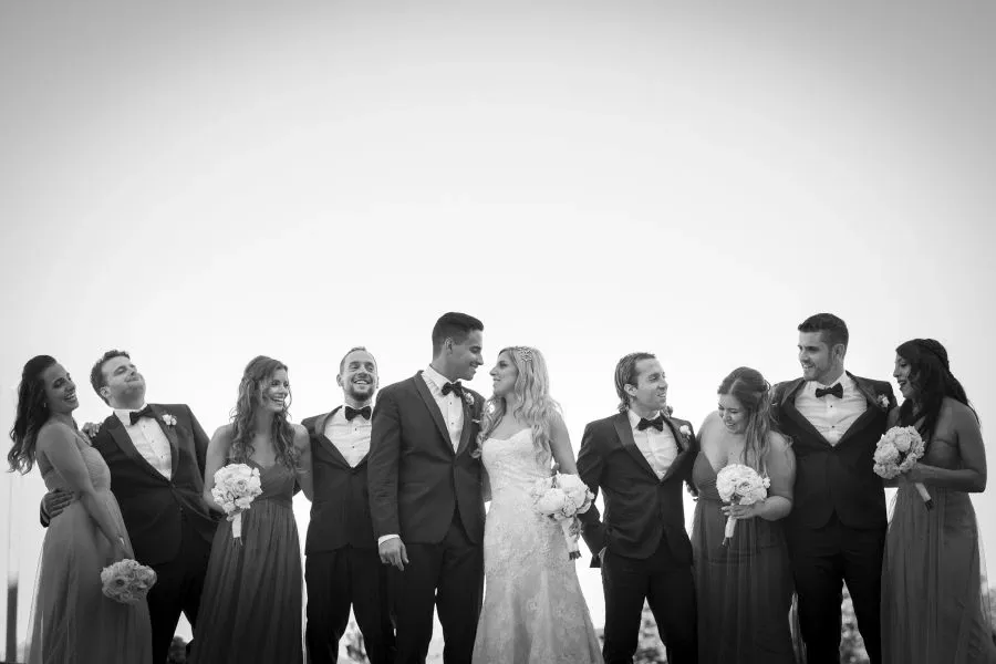A group of bridesmaids and groomsmen surrounds a joyful bride and groom in this wedding photography image by Foto Studio Franca Sorrenti. The bride, in a stunning lace gown, stands arm in arm with the groom, who is dressed in a classic tuxedo. The bridal party, elegantly attired in matching dresses and suits, holds bouquets and smiles warmly, embodying the celebratory and elegant atmosphere of the occasion. This image captures a candid moment of togetherness, highlighting the vendor's expertise in wedding photography.