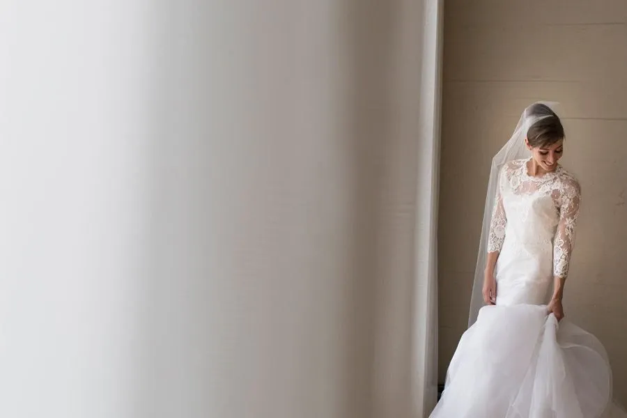 A bride stands gracefully in a softly lit room, captured by Foto Studio Franca Sorrenti, a vendor specializing in wedding photography. She wears an elegant, long-sleeved lace wedding gown with a delicately embroidered bodice and a flowing tulle skirt. The bride, looking down, adjusts the layers of her dress, revealing intricate details of the fabric. Her hair is styled in an elegant updo and she is adorned with a sheer veil. The neutral background emphasizes the simplicity and elegance of the moment, making it an ideal capture for a wedding day.