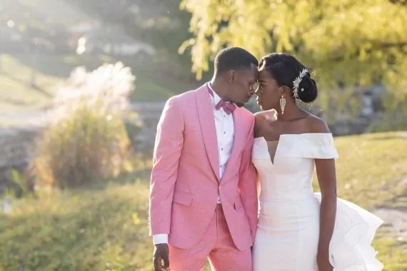 A newlywed couple is captured in an intimate moment, standing closely together in a serene outdoor setting. The groom wears a stylish pink suit paired with a white shirt and a matching pink bow tie, exuding a modern and fashionable vibe. The bride is elegantly dressed in an off-the-shoulder white wedding gown, featuring a long cascading bow at the back, and her hair is adorned with a delicate hairpiece. The background is beautifully dotted with lush greenery and gentle sunlight filtering through the trees, creating a romantic ambiance. This image is brought to life by Foto Studio Franca Sorrenti, specializing in Wedding Photography, as featured on Mariage Elegant, Canada.