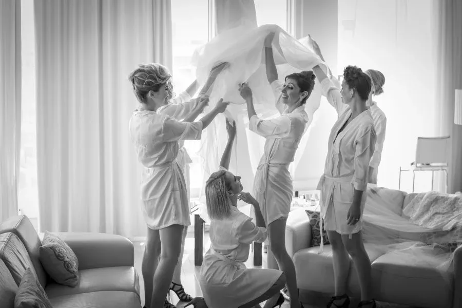 In this black and white image by Foto Studio Franca Sorrenti, a group of five women, likely the bride and her bridesmaids, are joyfully preparing for the wedding celebration. They are all dressed in elegant, matching robes, standing in a well-lit room with large windows. The women are holding up a sheer fabric, possibly a wedding veil or part of a dress, with smiles and laughter, capturing a candid and heartfelt moment. This scene exemplifies the expertise in wedding photography offered by the vendor, showcasing their ability to capture intimate, memorable moments in the bridal suite.