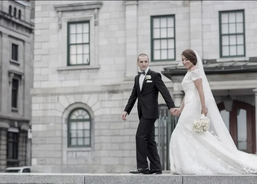 A newlywed couple elegantly poses outside a historic stone building, holding hands and walking gracefully. The bride is in a classic, white lace gown with a long, flowing train and veil, while the groom wears a tailored black tuxedo with a bow tie. The backdrop of the image features arched windows and detailed masonry, adding a timeless feel to the photo. This moment, captured by Foto Studio Franca Sorrenti, showcases their expertise in the wedding photography category, emphasizing elegance and tradition. This image is featured on Mariage Elegant, a wedding planning and vendor listing platform in Canada.
