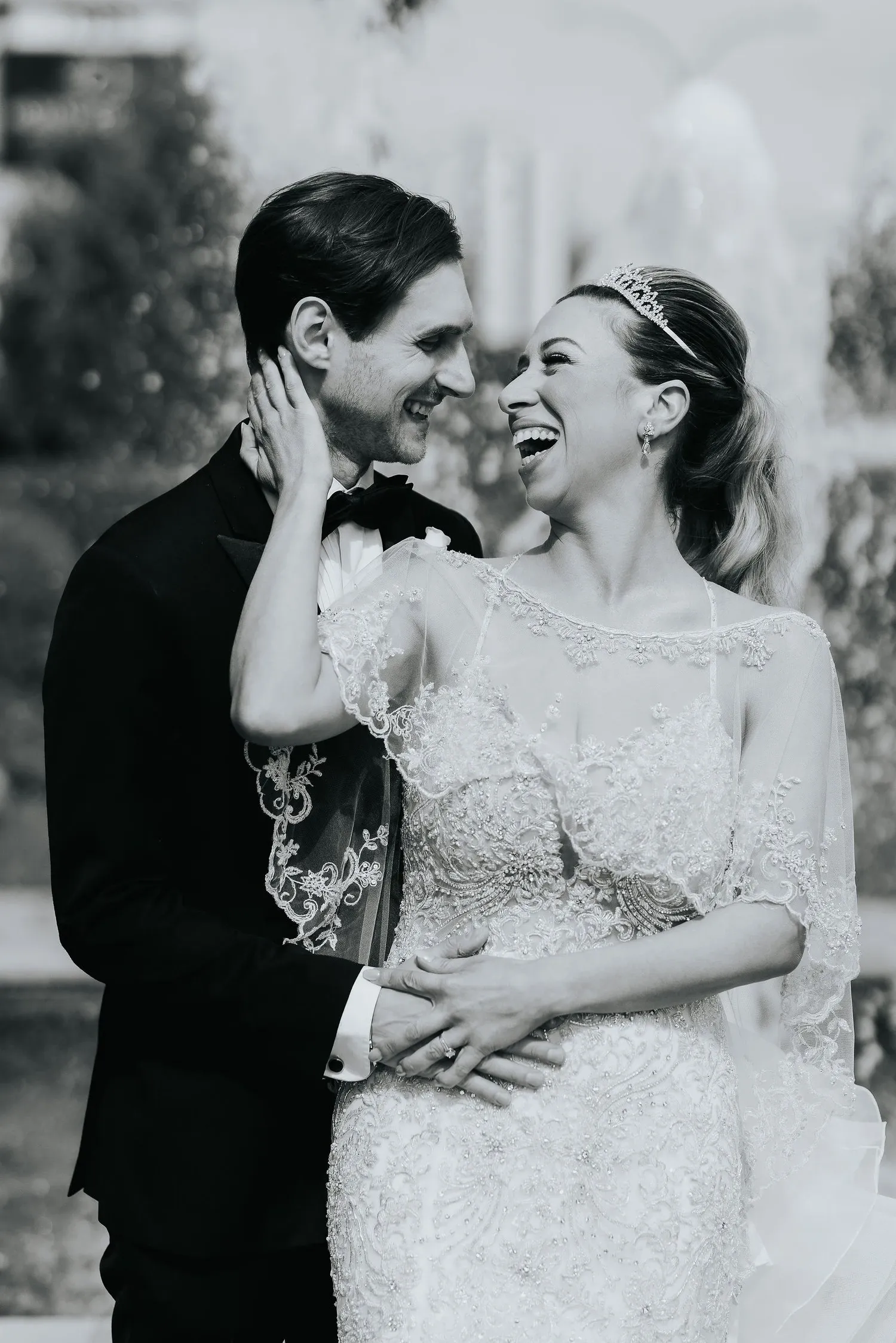 A joyful bride and groom stand close together in a beautiful outdoor setting, both dressed elegantly for their wedding day. The bride is wearing a stunning lace wedding gown with intricate detailing and a delicate veil, her hair styled beautifully with a tiara. The groom is in a classic black tuxedo with a bow tie. They are captured in a candid moment, smiling warmly at each other, exuding happiness and affection. The background suggests an elegant garden with a fountain, adding a romantic atmosphere. This image showcases the professional wedding photography by Braulio Rocha Photography, highlighting their expertise in capturing memorable, heartwarming moments in the wedding category.