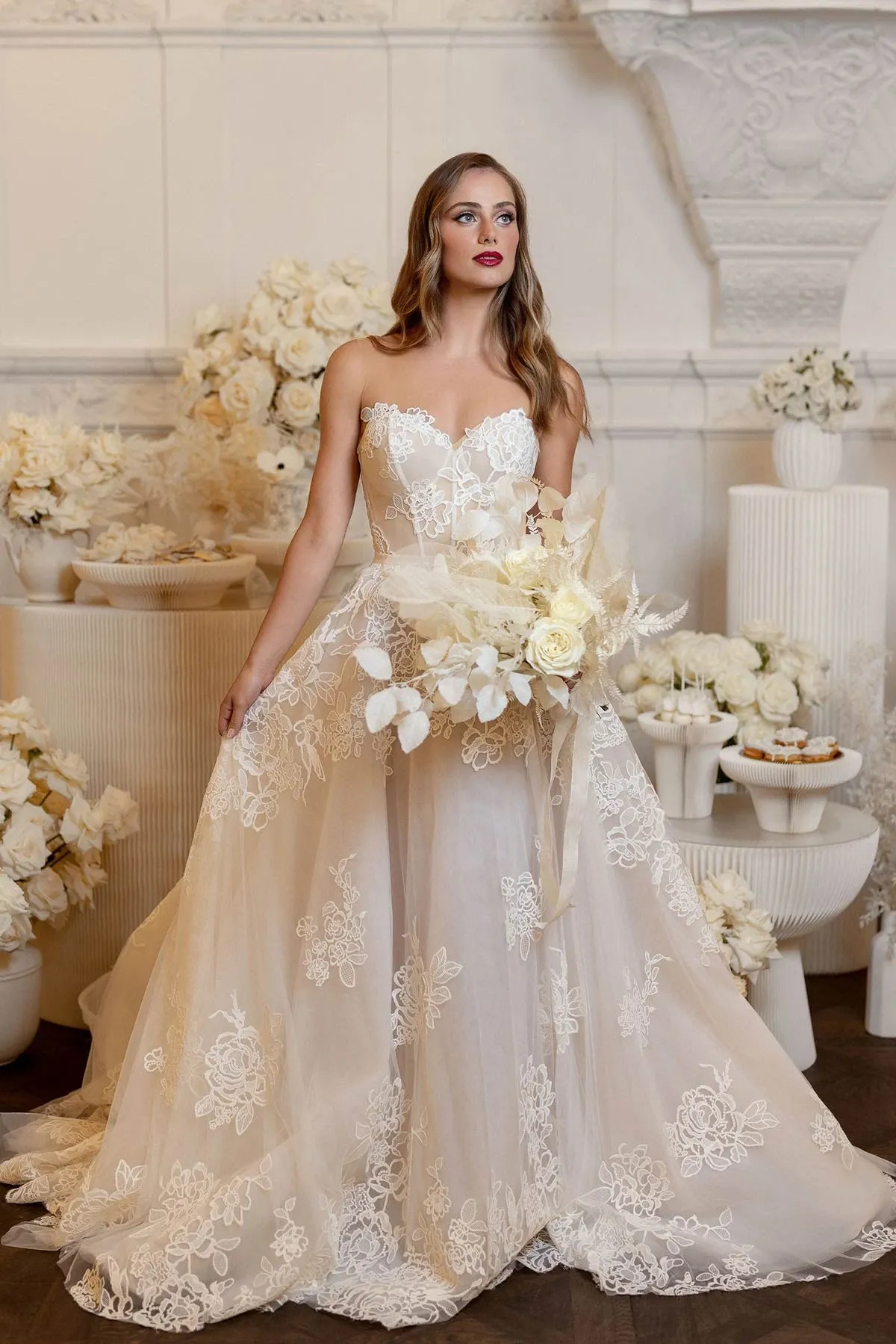 This image by Braulio Rocha Photography, a vendor in the Wedding Photography category, showcases a bride in an elegant wedding gown. The dress features intricate lace detailing with a strapless, sweetheart neckline. She holds a bouquet of white flowers, complementing the delicate floral patterns on her gown. The setting is adorned with white roses and soft-toned floral arrangements, creating a romantic and sophisticated ambience. The bride's poised expression and classic styling enhance the timeless elegance of this wedding scene, ideal for showcasing fine wedding photography.