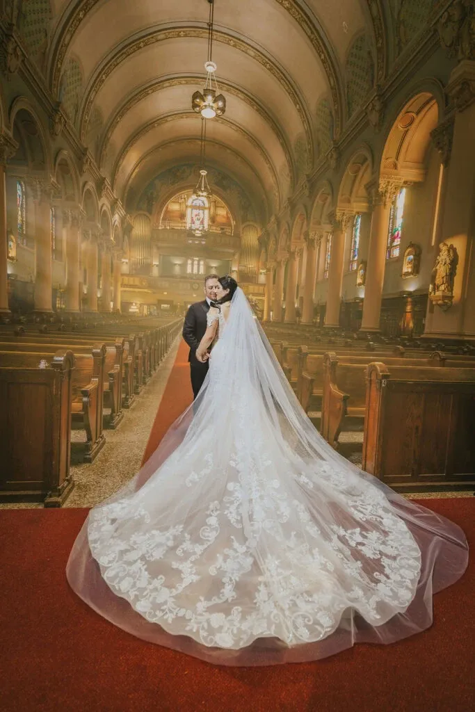 In this stunning image by Braulio Rocha Photography, a couple is captured in a romantic embrace at the altar of a grand church, underlining the wedding photography category. The bride's exquisite lace dress extends into a long, flowing train that gracefully spreads over the rich red carpet, creating a striking contrast. The church's intricate architecture, with its high arched ceilings and elegant stained glass windows, adds to the majestic atmosphere, highlighting the significance of the special occasion. The couple's intimate moment is beautifully framed by the warm, ambient lighting that filters through the ornate interior.