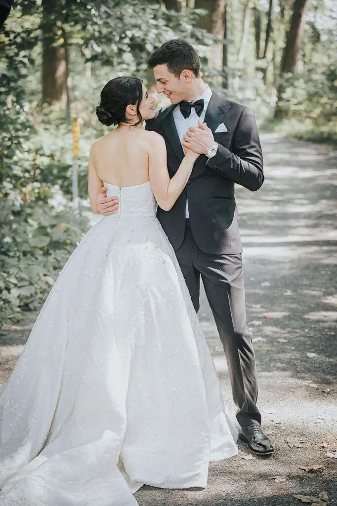 A bride and groom share an intimate moment on a wooded path. The bride is wearing an elegant, strapless white gown with intricate detailing, while the groom is dressed in a classic black tuxedo with a bow tie. They are gazing into each other's eyes as they hold hands, creating a romantic scene. This image captures a dreamy outdoor wedding setting, showcasing the photography style of Braulio Rocha Photography, a specialist in wedding photography featured on Mariage Elegant, Canada.