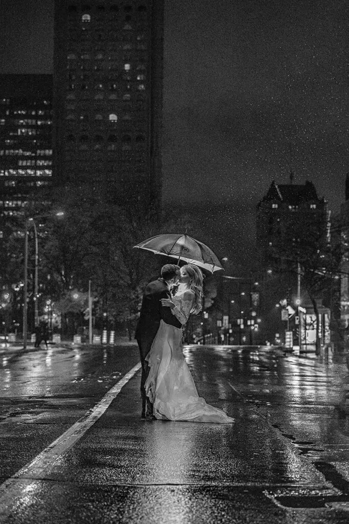 In this black and white image captured by Braulio Rocha Photography, a couple is seen embracing on a rainy city street at night. The bride, wearing a flowing wedding gown, and the groom, in a classic suit, stand under a transparent umbrella amidst glistening wet pavement. The backdrop features tall buildings with lit windows, adding an urban and romantic feel to the wedding photography. This image beautifully encapsulates the intimate, timeless atmosphere suitable for Mariage Elegant's wedding planning and vendor listing in Canada.