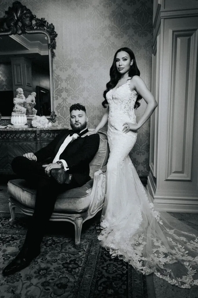 This elegant black-and-white image by Braulio Rocha Photography, a vendor specializing in wedding photography, features a bride and groom in a luxurious, vintage setting. The bride stands gracefully, showcasing her intricately embroidered, form-fitting wedding gown with a long lace train. Her hair is styled in soft waves, enhancing her refined look. The groom, dressed in a classic tuxedo, sits confidently on an ornate chair, exuding sophistication. The backdrop includes an antique dresser with decorative elements like porcelain and a large, ornate mirror, adding to the timeless elegance of the scene, which is perfect for wedding inspiration.