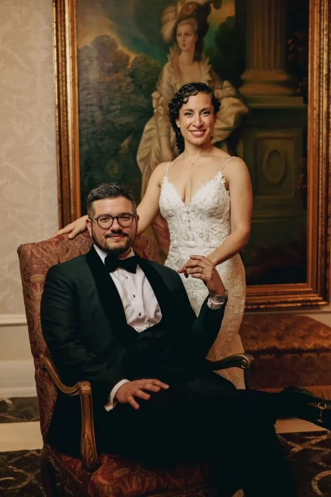 A bride and groom are elegantly posed indoors, captured by Braulio Rocha Photography, a vendor in the Wedding Photography category. The groom, seated in a classic armchair, wears a black tuxedo with a white dress shirt and bow tie. The bride stands beside him in a lace wedding gown, gently resting her hand on the groom's shoulder. Both are smiling softly at the camera. Behind them is a large, ornate painting, adding a touch of sophistication to the setting. The image reflects a classic and refined wedding atmosphere, perfect for showcasing high-end wedding photography services.