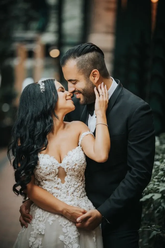 A joyful couple, elegantly dressed in wedding attire, shares an intimate moment in an outdoor setting. The bride, wearing a lace-embellished off-shoulder gown with intricate floral details, lovingly places her hand on the groom's cheek. Her long, dark hair cascades down, adorned with a delicate headpiece. The groom, in a classic black suit, embraces her with a warm smile. This image showcases the romantic and candid photography style of Braulio Rocha Photography, a vendor specializing in Wedding Photography for Mariage Elegant, Canada. Lush greenery in the background adds to the serene and romantic atmosphere.