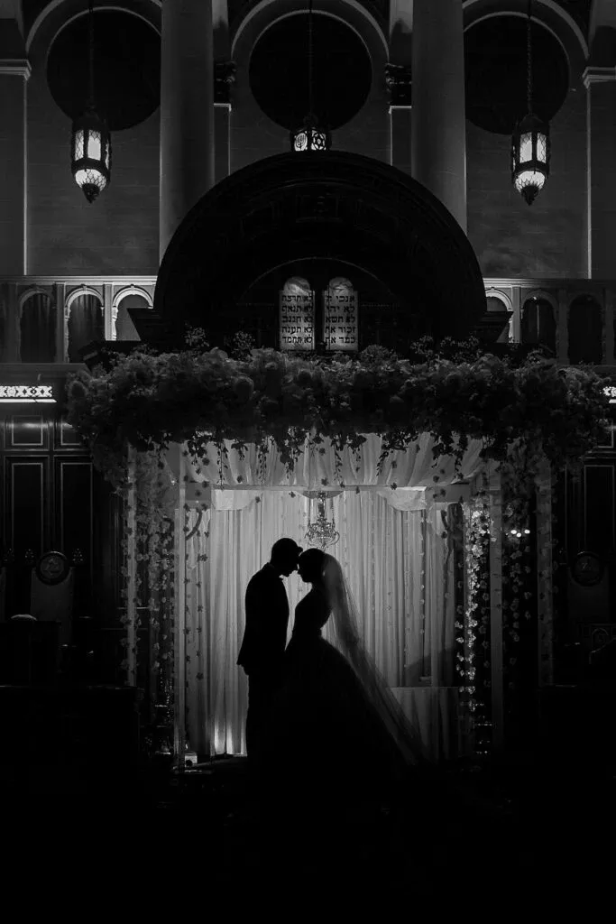 This image by Braulio Rocha Photography, showcased in the Wedding Photography category on Mariage Elegant, Canada, captures a striking black and white silhouette of a bride and groom sharing an intimate moment under a beautifully decorated wedding altar. The altar is adorned with cascading floral arrangements and illuminated softly, adding an ethereal glow to the scene. In the background, elegant architectural details and ornate hanging lanterns create a romantic and timeless atmosphere, perfectly encapsulating the essence of a dream wedding.