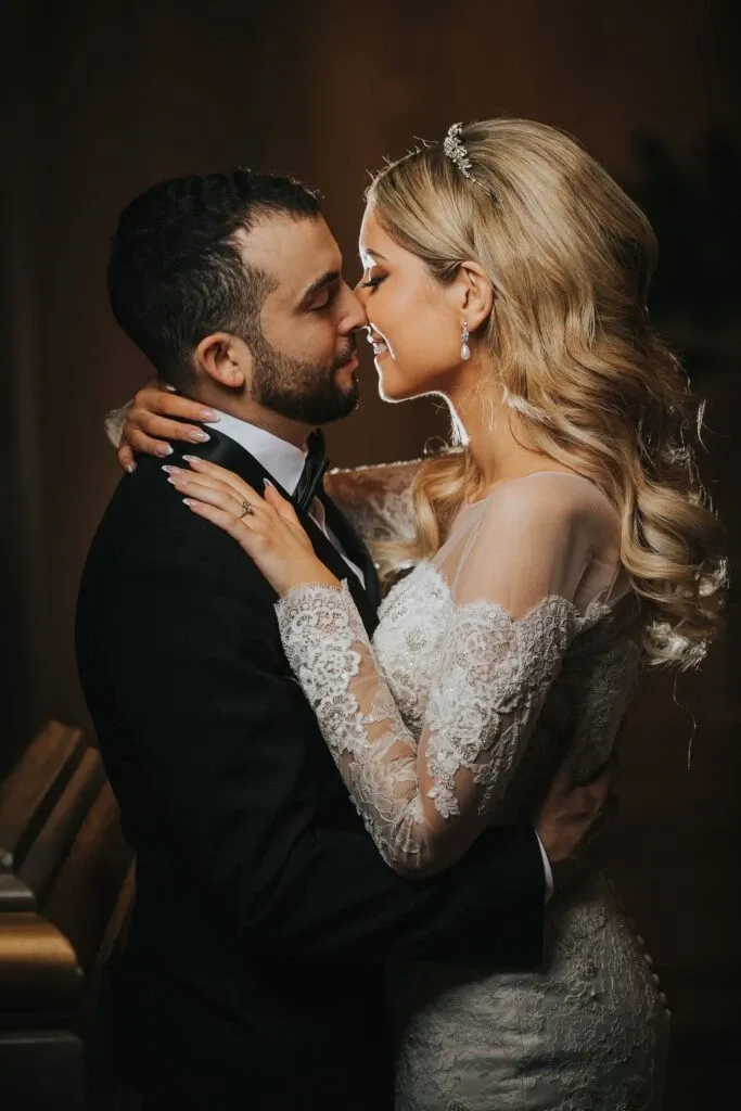 A bride and groom share a tender moment, embracing each other closely. She has long, wavy blonde hair adorned with a delicate tiara and wears an elegant, lace long-sleeve wedding gown. The groom is dressed in a classic black tuxedo with a white shirt and bow tie. They are intimately leaning towards each other, eyes closed, with warm expressions of love. The soft lighting casts a romantic glow over them, highlighting the intricate details of the bride's dress. This image is captured by Braulio Rocha Photography, a vendor specializing in wedding photography for Mariage Elegant, Canada.