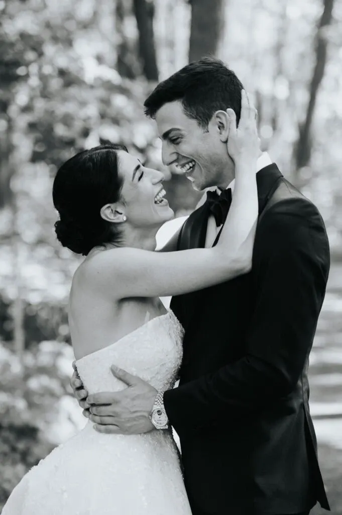 A joyful couple is captured in an intimate embrace in this black and white image by Braulio Rocha Photography, a vendor specializing in wedding photography. The bride, dressed in an elegant strapless gown, lovingly holds the groom's face with one hand while the other rests on his back. The groom, wearing a sharp suit, gazes affectionately into her eyes, their smiles radiant with happiness. The background features a serene forest setting, with sunlight filtering through the trees, enhancing the romantic atmosphere of this special moment.